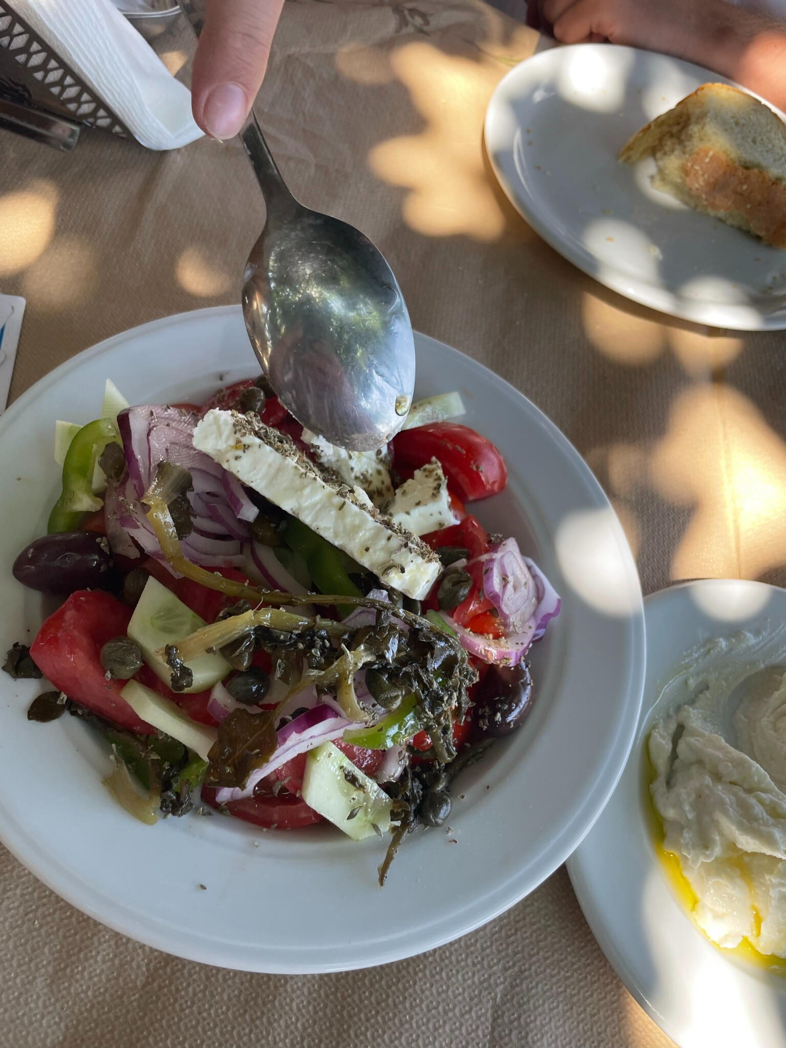 "A traditional Greek salad in Tinos, Greece, with fresh tomatoes, cucumbers, feta cheese, olives, and capers, drizzled with olive oil and served with bread and dip."