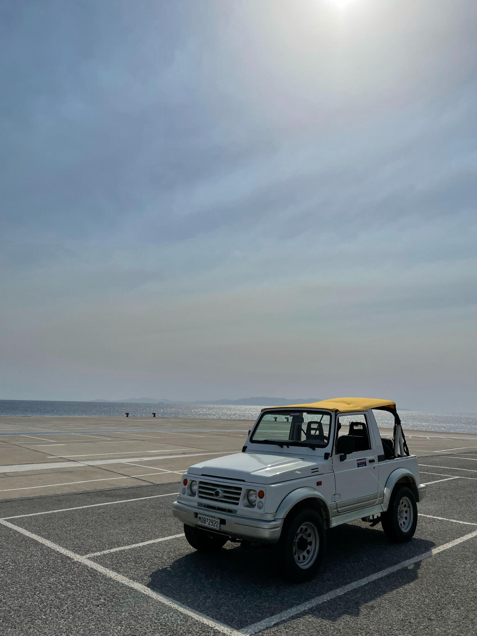 A classic white Suzuki jeep with a yellow soft top parked near the harbour on Tinos Island, Greece. The Aegean Sea glistens in the background under a clear blue sky, capturing the essence of slow, off-road travel and adventure on this Cycladic island.