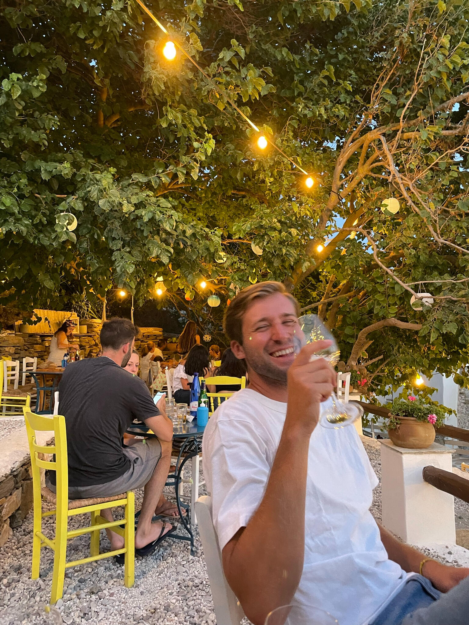 "A cheerful evening at a Greek taverna in Triantaros, Tinos. A man in a white shirt enjoys wine under glowing string lights, surrounded by rustic decor, yellow chairs, and lush greenery."