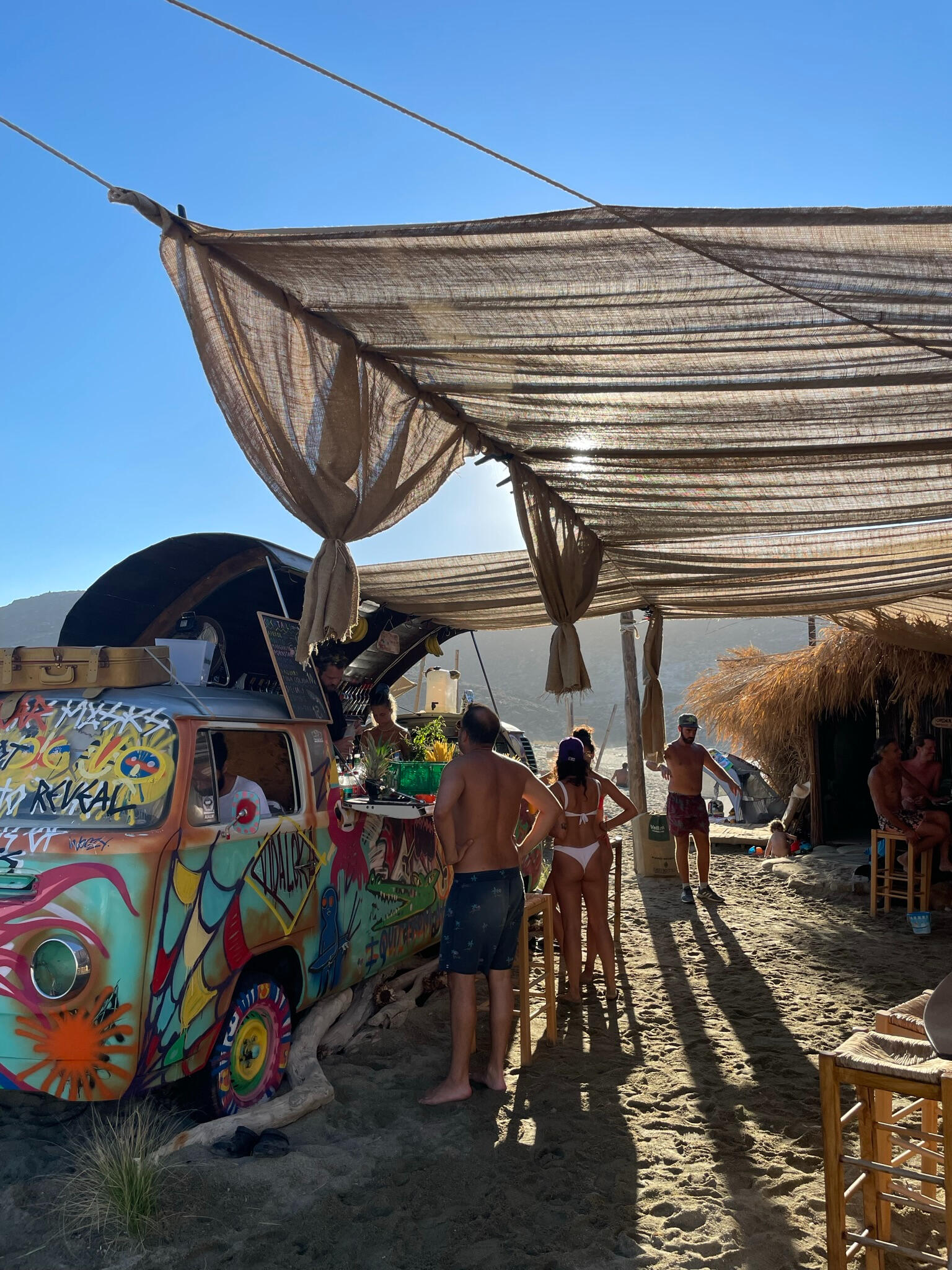 "A colorful vintage van-turned-beach bar on a sandy shore in Tinos, Greece. Guests enjoy drinks under a shaded canopy, creating a laid-back island vibe."