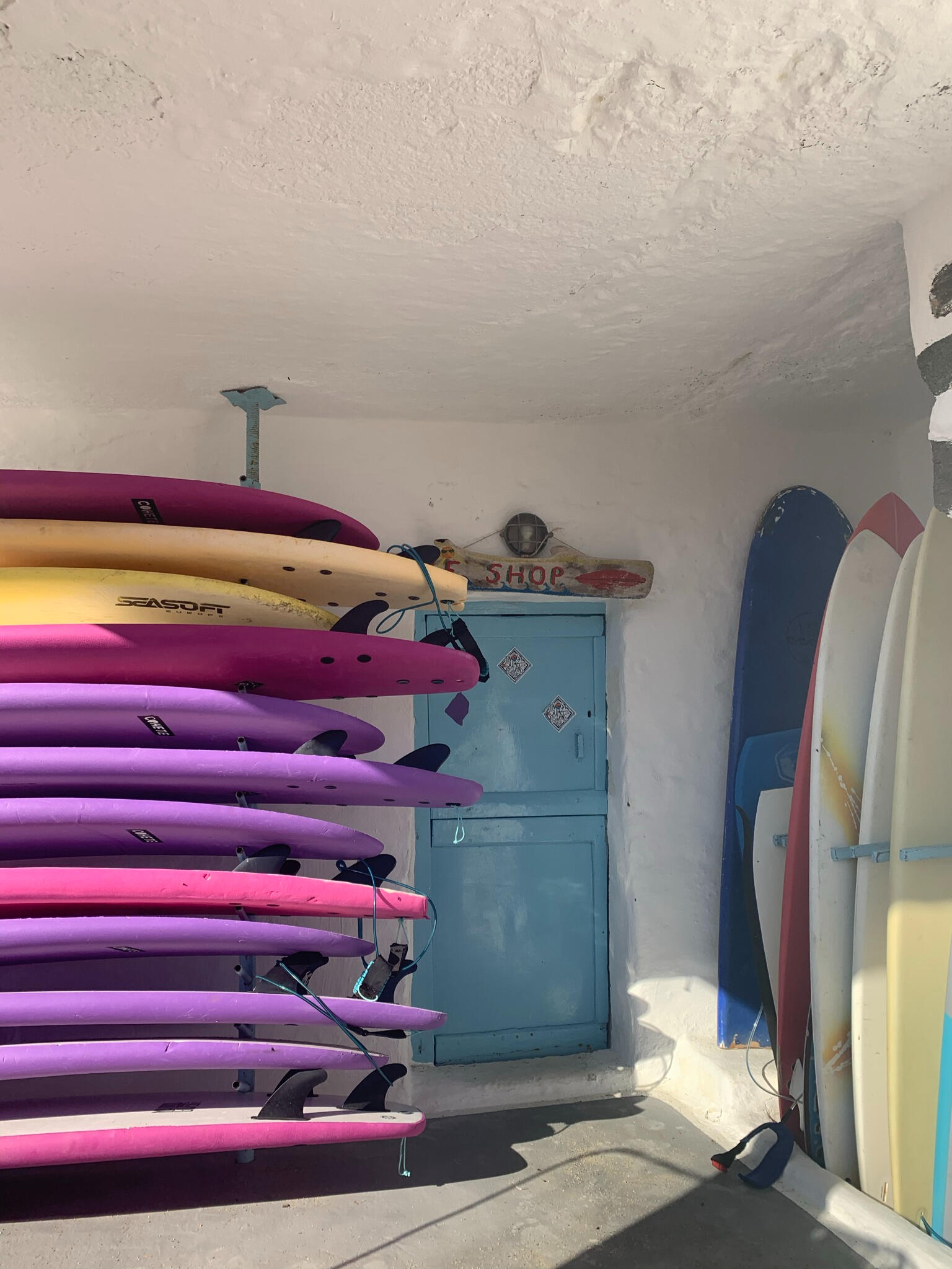 "Colorful surfboards and kayaks stacked outside a small surf shop on Tinos Island, Greece. The rustic blue door and Cycladic walls reflect the island’s beach culture and adventure vibe."