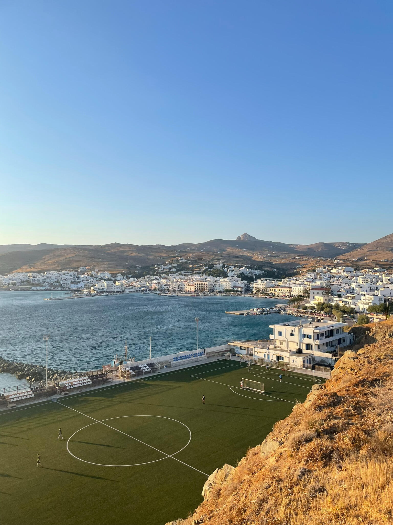 "Scenic view of a football pitch in Tinos Town, Greece, overlooking the Aegean Sea. The setting sun casts a golden light on the Cycladic landscape, with whitewashed buildings and rolling hills in the background, showcasing the charm of island life."