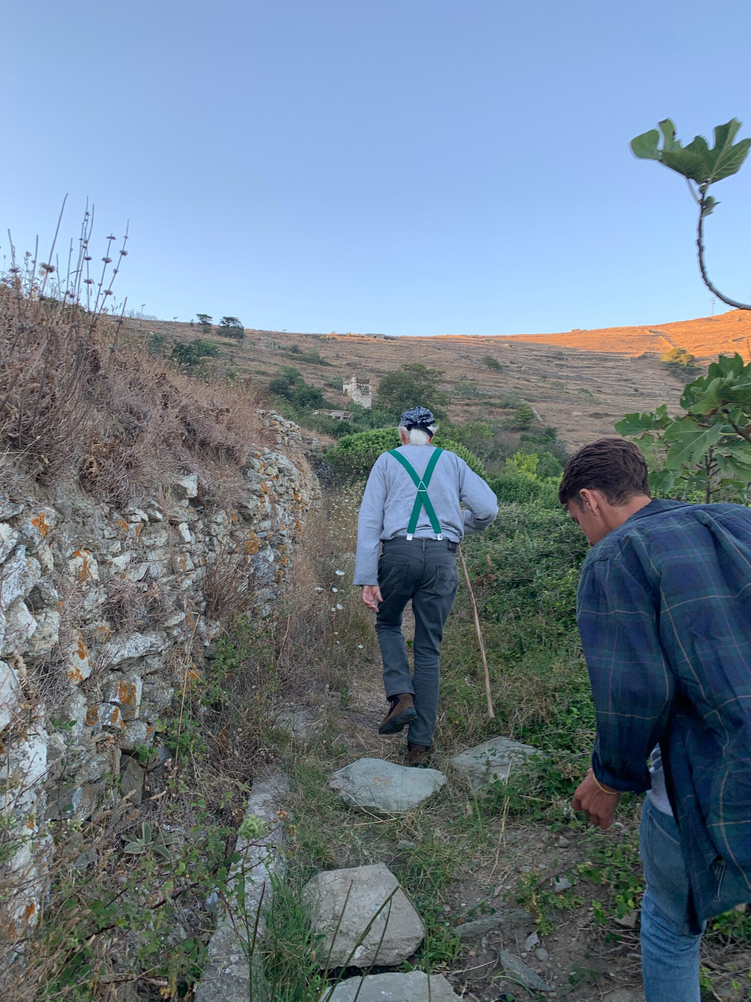 Two men following an elderly local guide with green suspenders on a rugged hiking trail in Tinos, Greece. The path winds through a dry stone wall and hillside vegetation, offering an authentic experience of rural Cycladic landscapes and nature exploration.