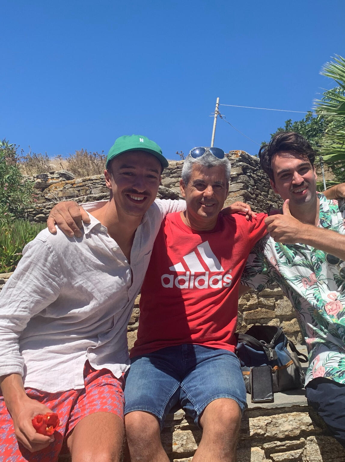 Three men smiling and sitting together under the bright Greek sun in Tinos, Greece. One wears a green cap, another a red Adidas shirt, and the third a floral shirt, symbolizing local hospitality, Greek island friendships, and authentic cultural experiences