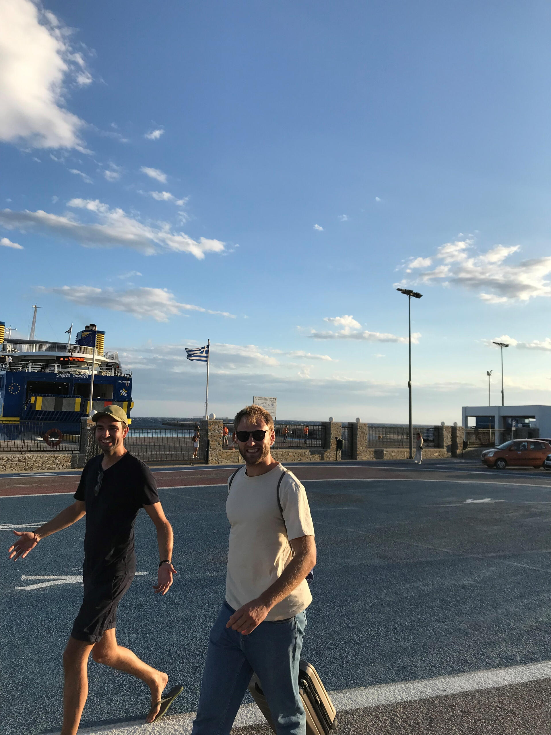 "Two men walk and smile near a ferry port in Greece. One carries a suitcase, and a blue and yellow ferry with a Greek flag is docked behind them. The warm evening light creates a relaxed vibe."