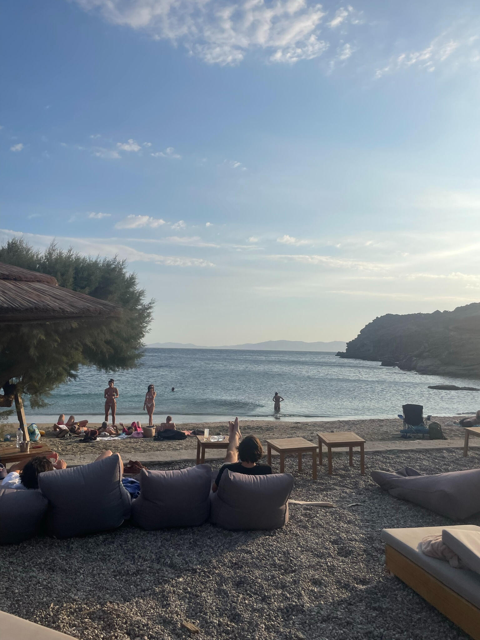 "Relaxing evening at Bianco Beach, Tinos, Greece. Guests lounge on cushions by the Aegean Sea, enjoying the sunset, crystal-clear waters, and peaceful island vibes."