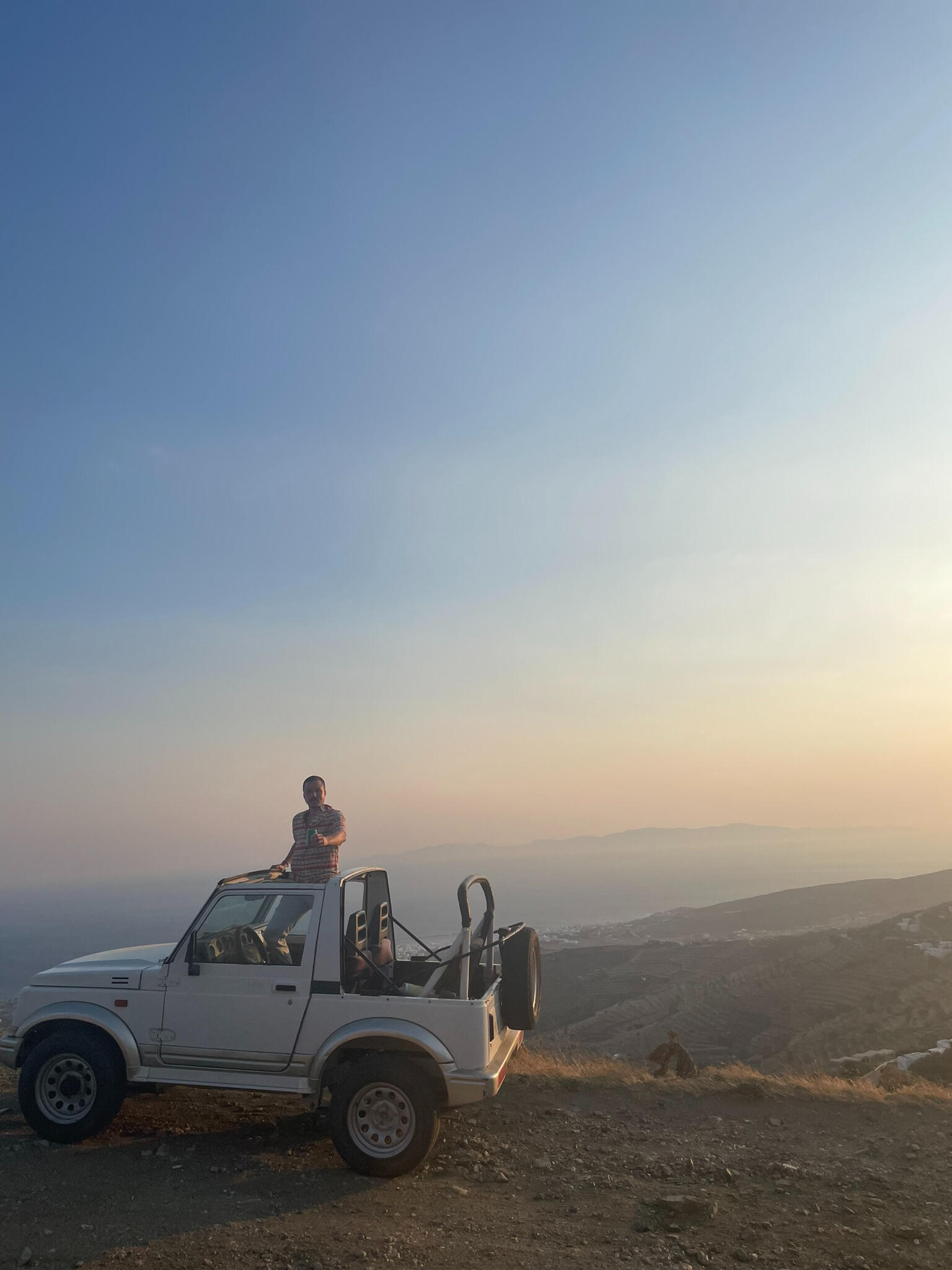 "A man stands on a white Suzuki Jimny 4×4 at sunset in Tinos, Greece. Overlooking vast Cycladic landscapes, the scene captures adventure and off-road island exploration."