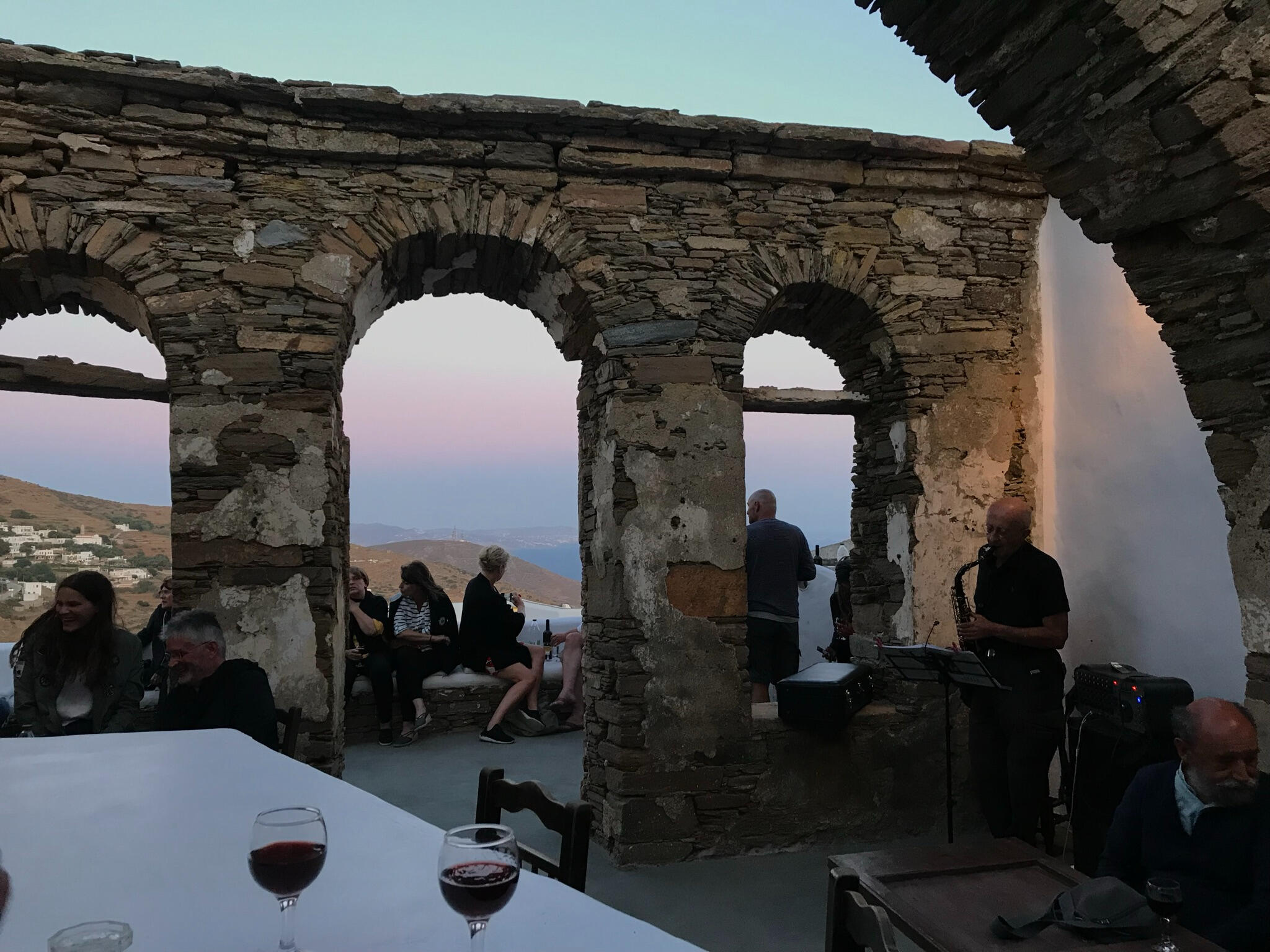 "Sunset at Bellis Winery in Arnados, Tinos, Greece. Guests enjoy wine, live saxophone music, and panoramic Cycladic views through ancient stone arches in a magical setting."