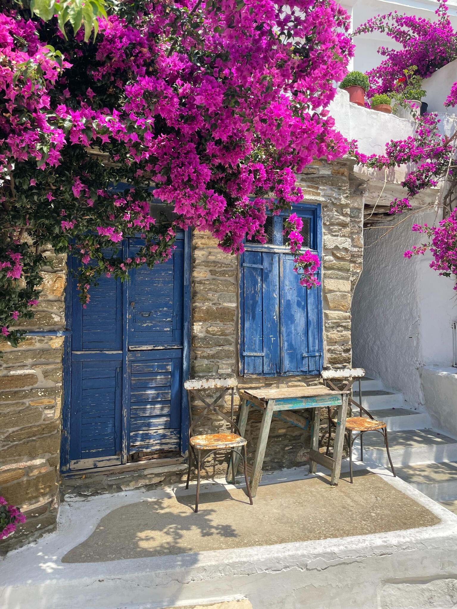 "A traditional stone house in Dyo Choria, Tinos, Greece, with blue wooden doors and vibrant bougainvillea. A picturesque corner capturing Cycladic charm and village life."