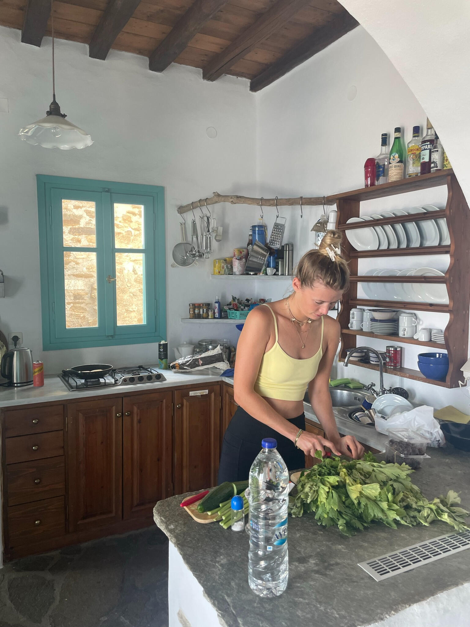 "A woman prepares fresh ingredients in a traditional Greek kitchen on Tinos Island. Sunlight streams through blue shutters, adding warmth to the rustic setting."