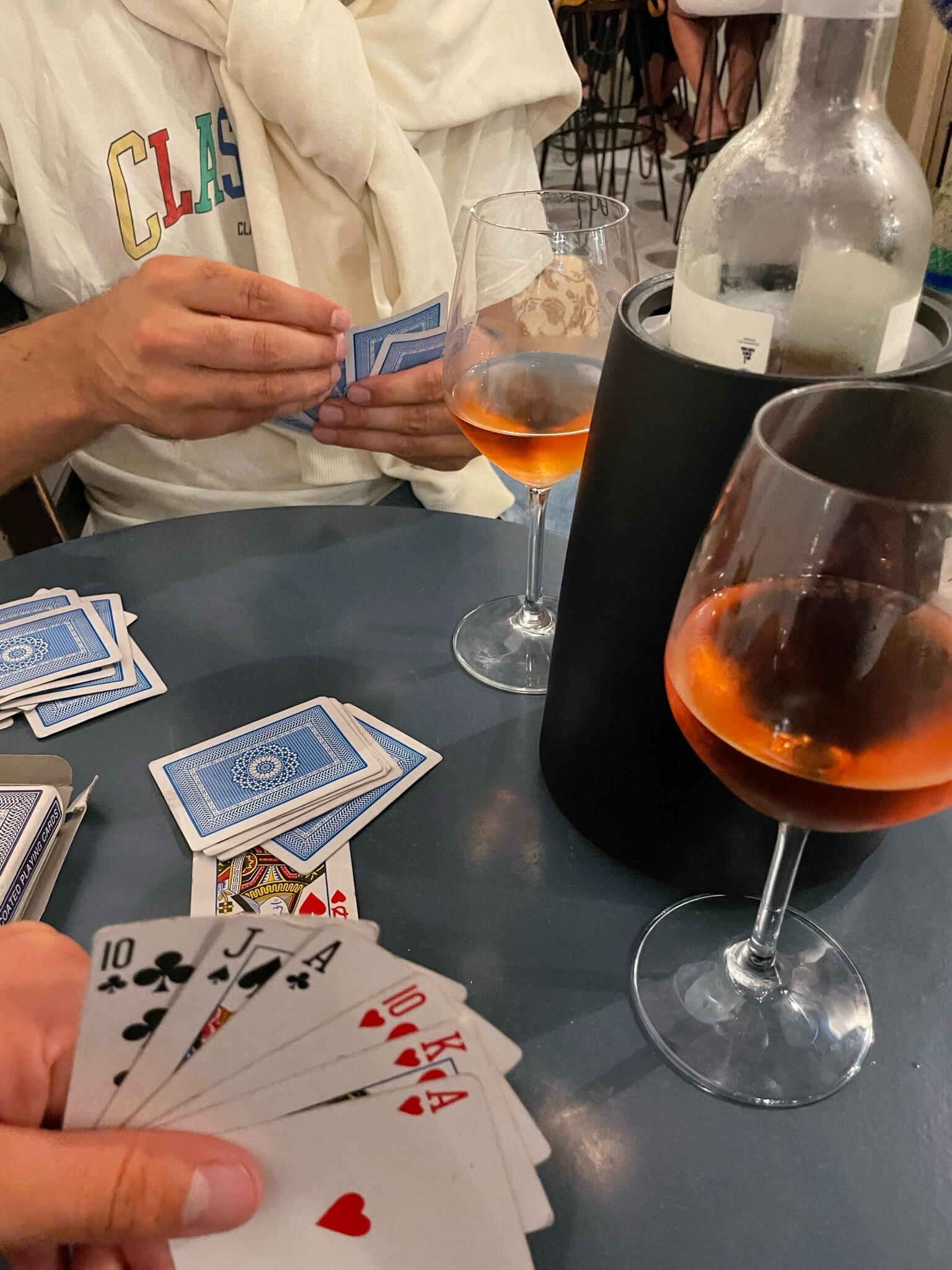 "A cozy evening at Antilalos in Tinos, Greece, with friends playing cards over glasses of rosé wine. A relaxed social atmosphere in a charming island café."